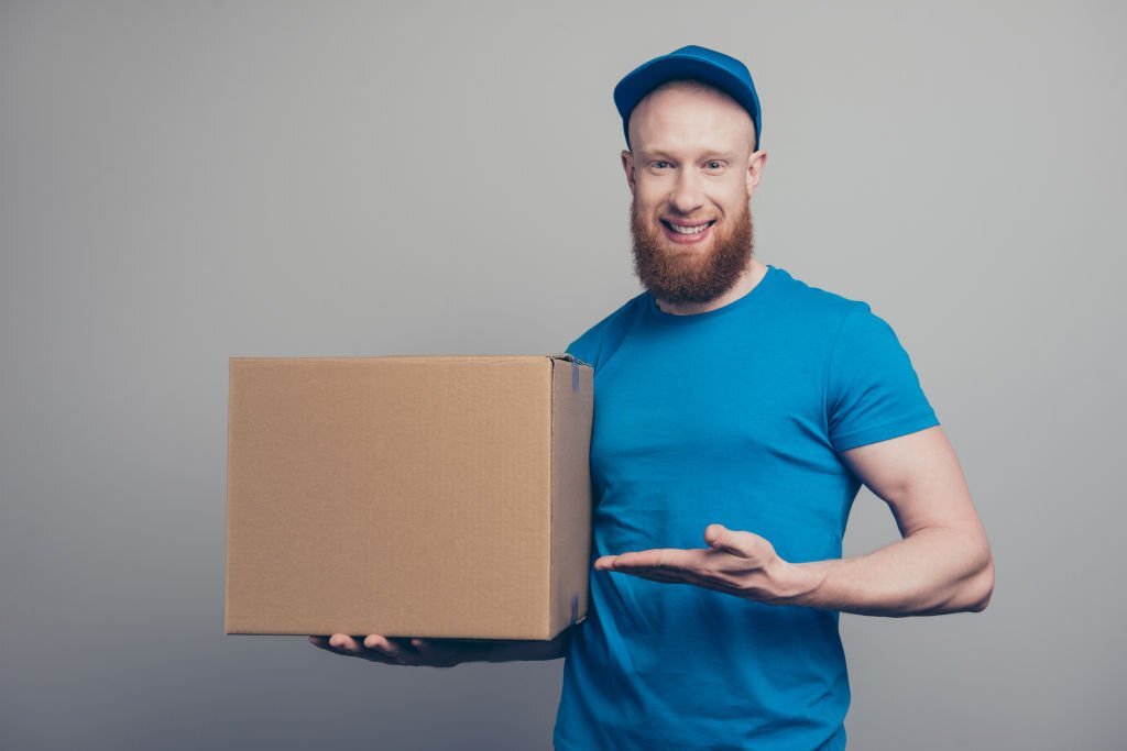 Portrait of nice handsome attractive cheerful positive bearded deliver in blue uniform holding in hands showing big carton box isolated over gray pastel background