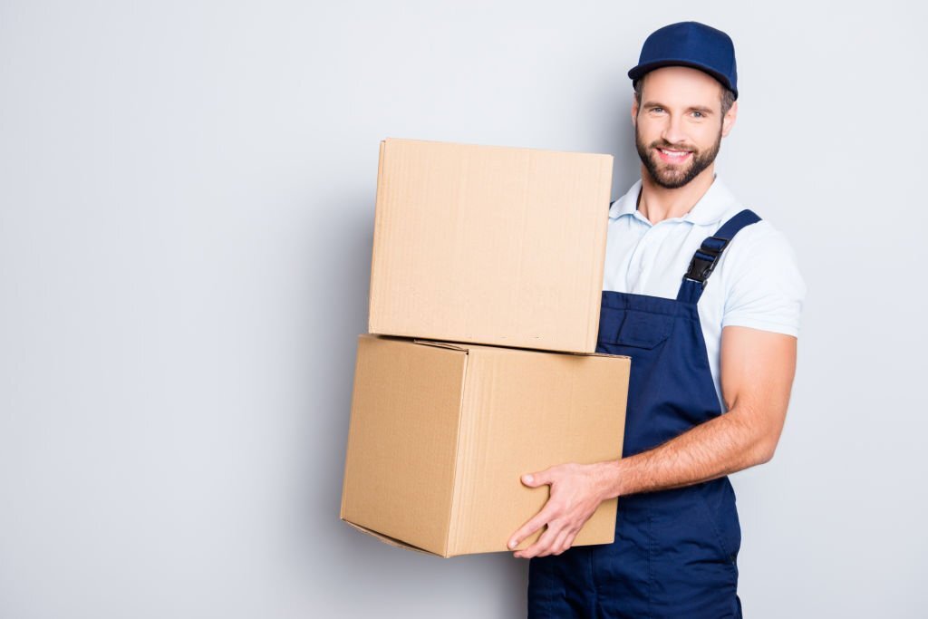 Portrait of attractive cheerful deliver in blue uniform with stubble looking at camera having two big boxes in hands, isolated on grey background