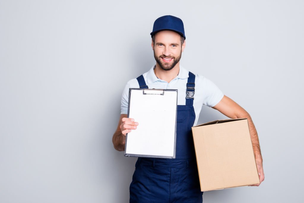 Portrait with empty place, copyspace of cheerful, attractive man having, carrying box in hands demonstrate clean paper in clipboard, looking at camera, isolated on grey background